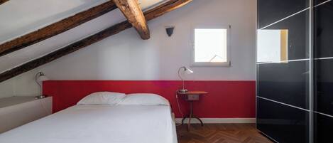 A bedroom with a sloped ceiling, a bed, a bedside table, a lamp, a wardrobe, and a window.