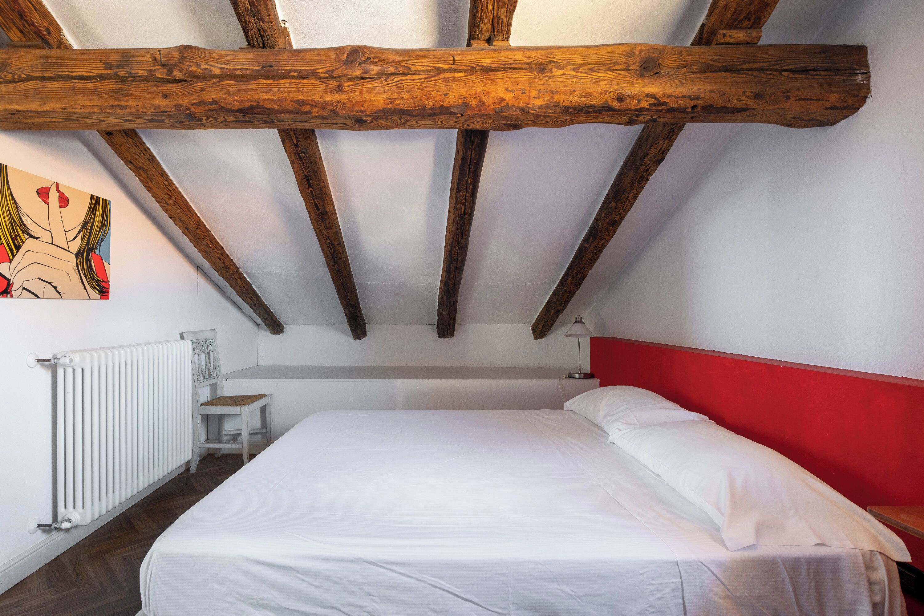 A bedroom with a wooden ceiling, white walls, a red accent wall, a white bed, and a radiator.