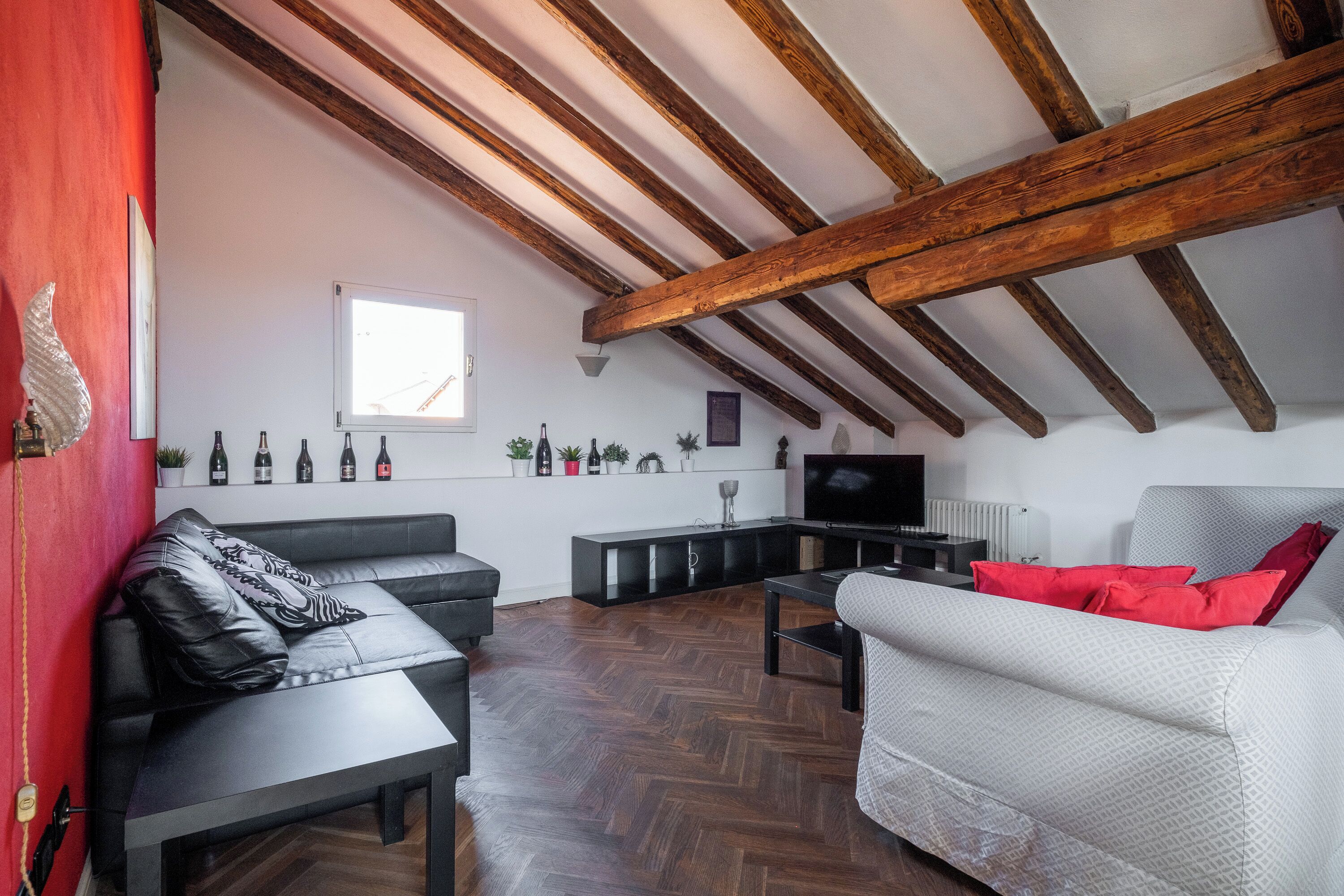 Uma sala de estar com piso de madeira em padrão herringbone, um sofá de couro preto, um sofá cinza com almofadas vermelhas, uma mesa de centro preta e uma televisão em um suporte preto.