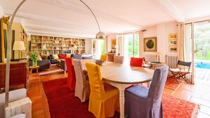 Ein geräumiges Zimmer mit einem großen runden Tisch, mehreren Stühlen, einem Bücherregal und Blick ins Freie.