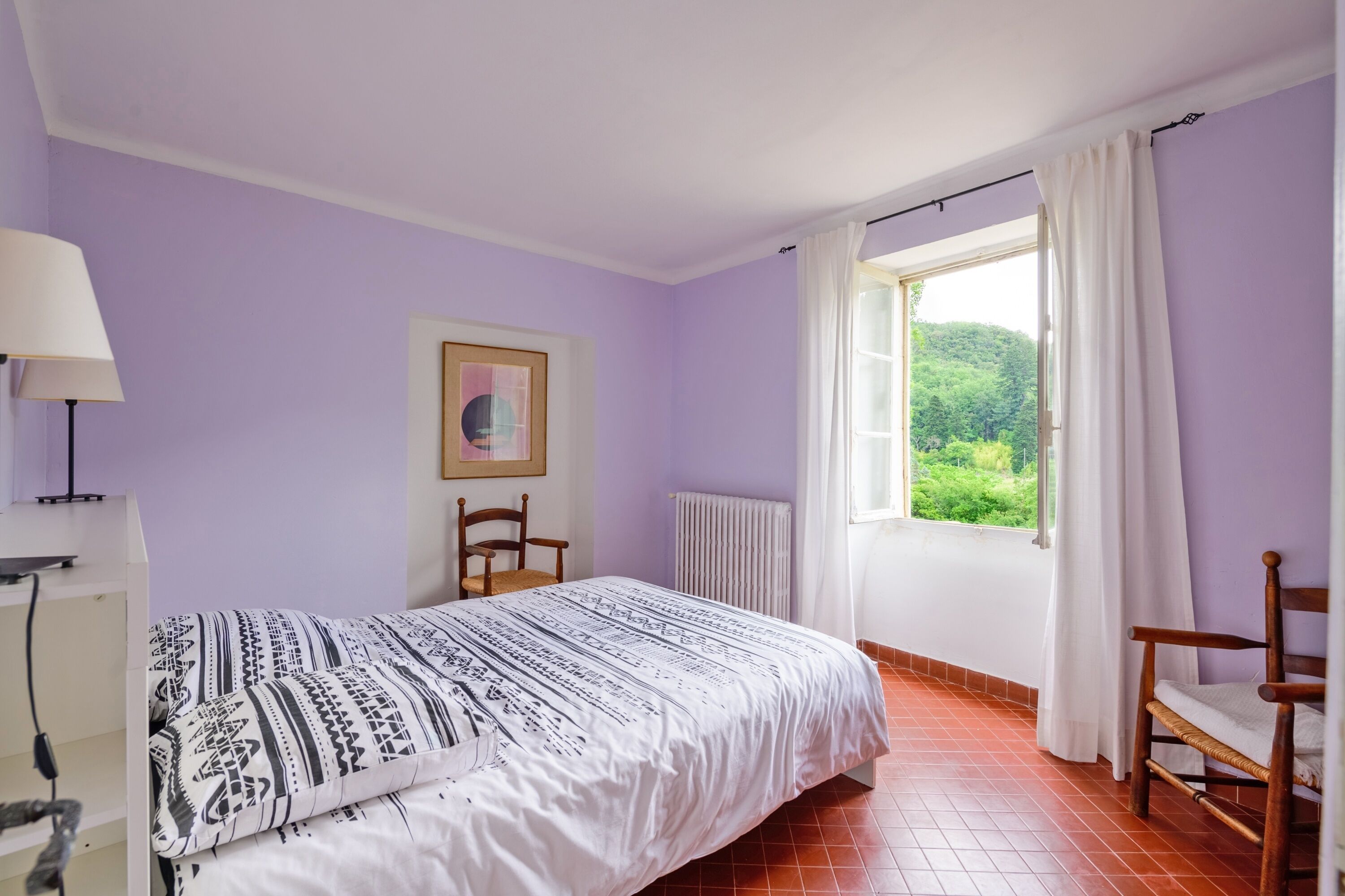 Un dormitorio con cama, una silla, una lámpara, un radiador y una ventana con cortinas.