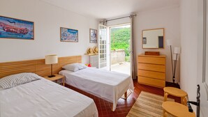 Ein Hotelzimmer mit zwei Betten, einem Holzkoffer, einem Spiegel und Blick auf eine grüne Landschaft durch eine offene Tür.