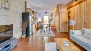 A modern living room with a TV, sofa, and coffee table. There is a kitchen area with a dining table and chairs. The room has wooden walls and a stone accent wall.