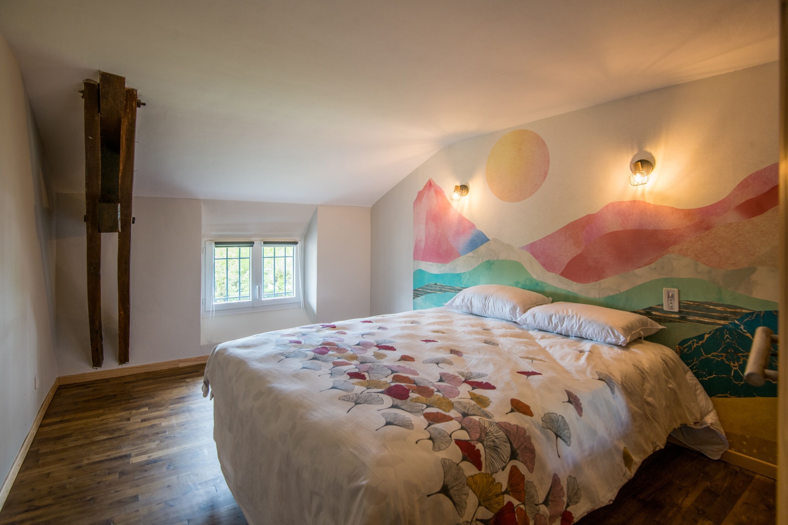 Une chambre avec un lit, une fenêtre, une échelle en bois et une fresque colorée sur le mur.