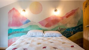 Quarto com cama, almofadas, um mural na parede e duas luzes fixadas na parede.