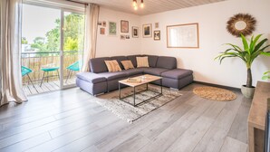 Una sala de estar moderna con un sofá seccional gris, una mesa de centro de madera y un amplio ventanal corredizo que da a un área exterior con terraza de madera y sillas azules.