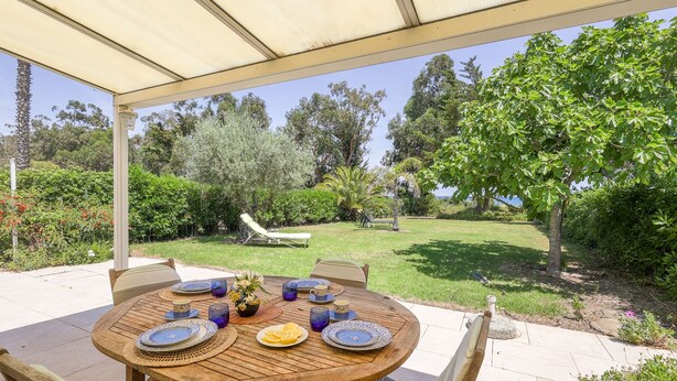 Una terrazza esterna con un tavolo rotondo in legno apparecchiata per mangiare, circondata da sedie, e una zona riparata con vista su un giardino rigoglioso e alberi.