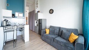 Un espace de vie moderne comprenant un canapé gris, deux coussins jaunes, un coin cuisine avec un réfrigérateur, un four à micro-ondes et une bouilloire, ainsi qu’un petit coin bar avec des tabourets.