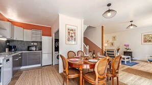 Una cocina moderna con armarios grises, horno empotrado y microondas. Hay un comedor con mesa y sillas de madera, y una escalera que conduce a otro nivel.