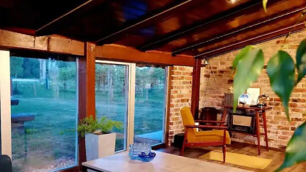 A cozy room with a wooden floor, a large window, a sofa, a table, and a brick wall.