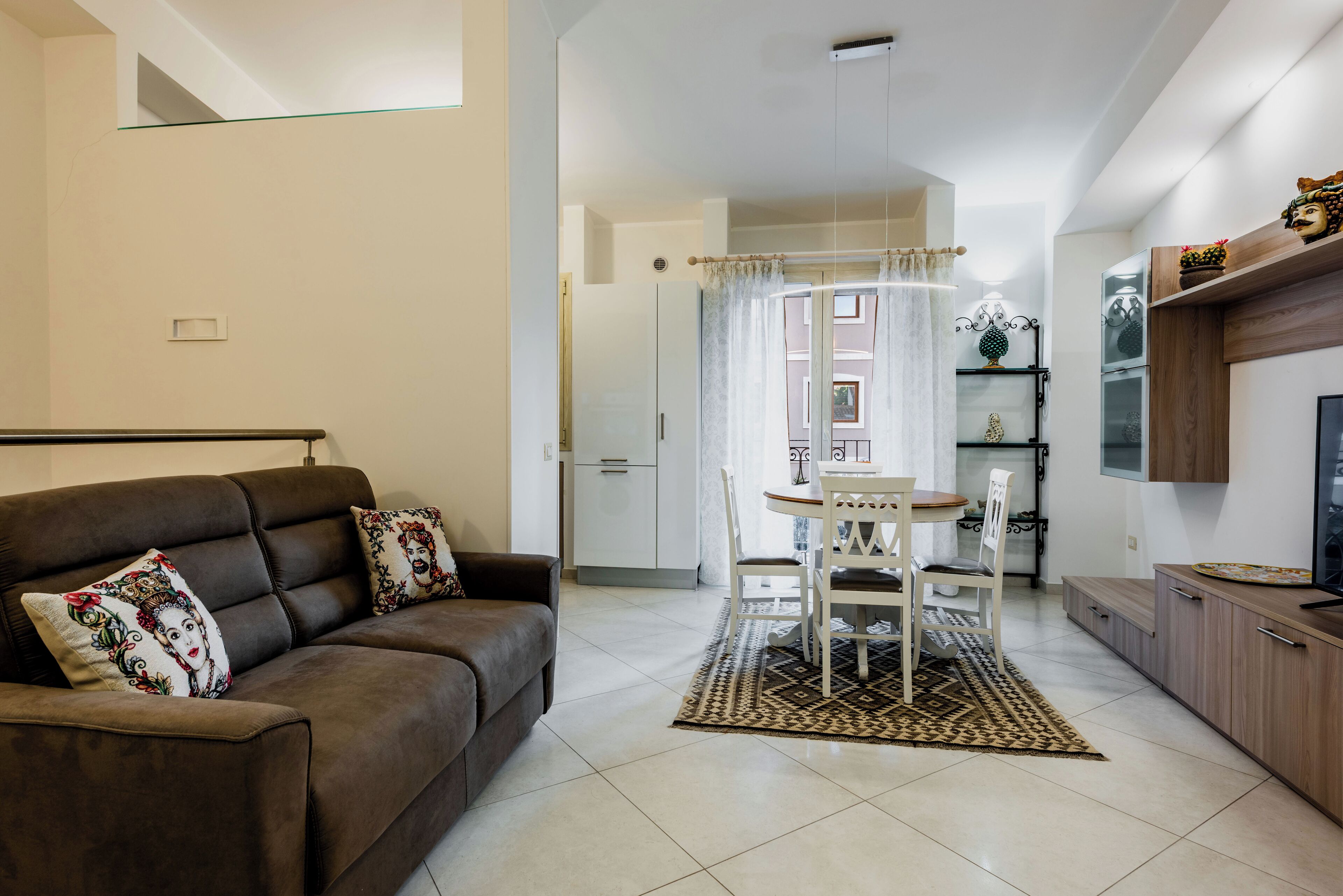 A living room with a brown sofa, a dining table with chairs, a kitchen area, and a TV on a wooden cabinet.
