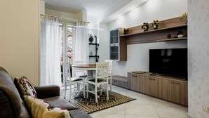 A modern living room with a brown leather sofa, a dining table with white chairs, a TV on a wooden cabinet, and a patterned rug.