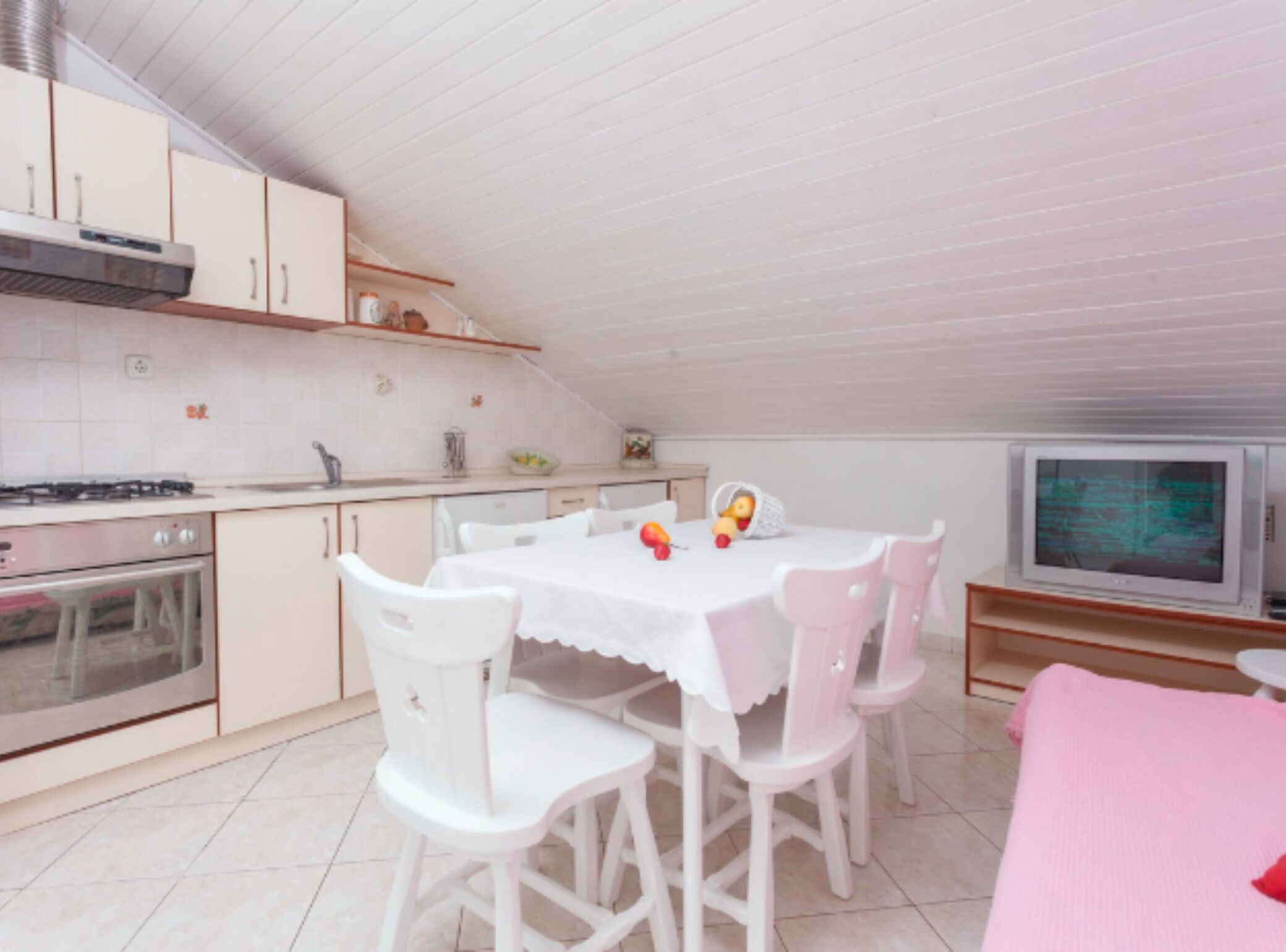 Cocina compacta con muebles blancos, estufa a gas y microondas. Hay una mesa de comedor con sillas blancas y un sofá rosa en la sala.