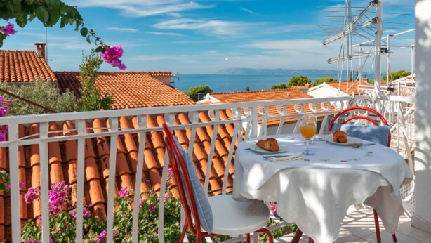 A terrace with a white table set for two, overlooking a coastal town with terracotta roofs. - Apartments Lili (Podgora)