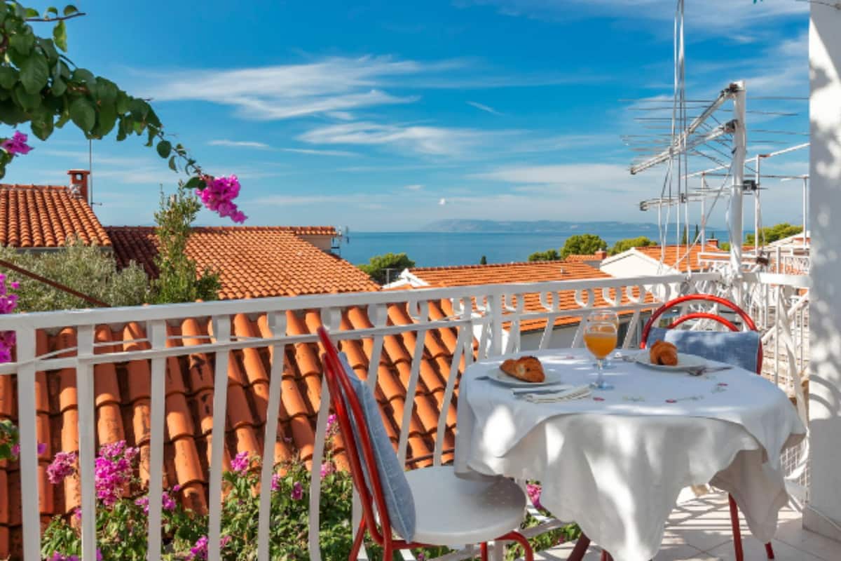 A terrace with a white table set for two, overlooking a coastal town with terracotta roofs.