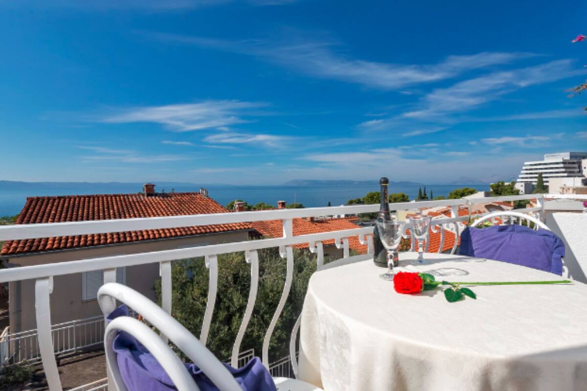 A terrace with a table set for two, overlooking a coastal view with red-tiled roofs and a clear blue sky.