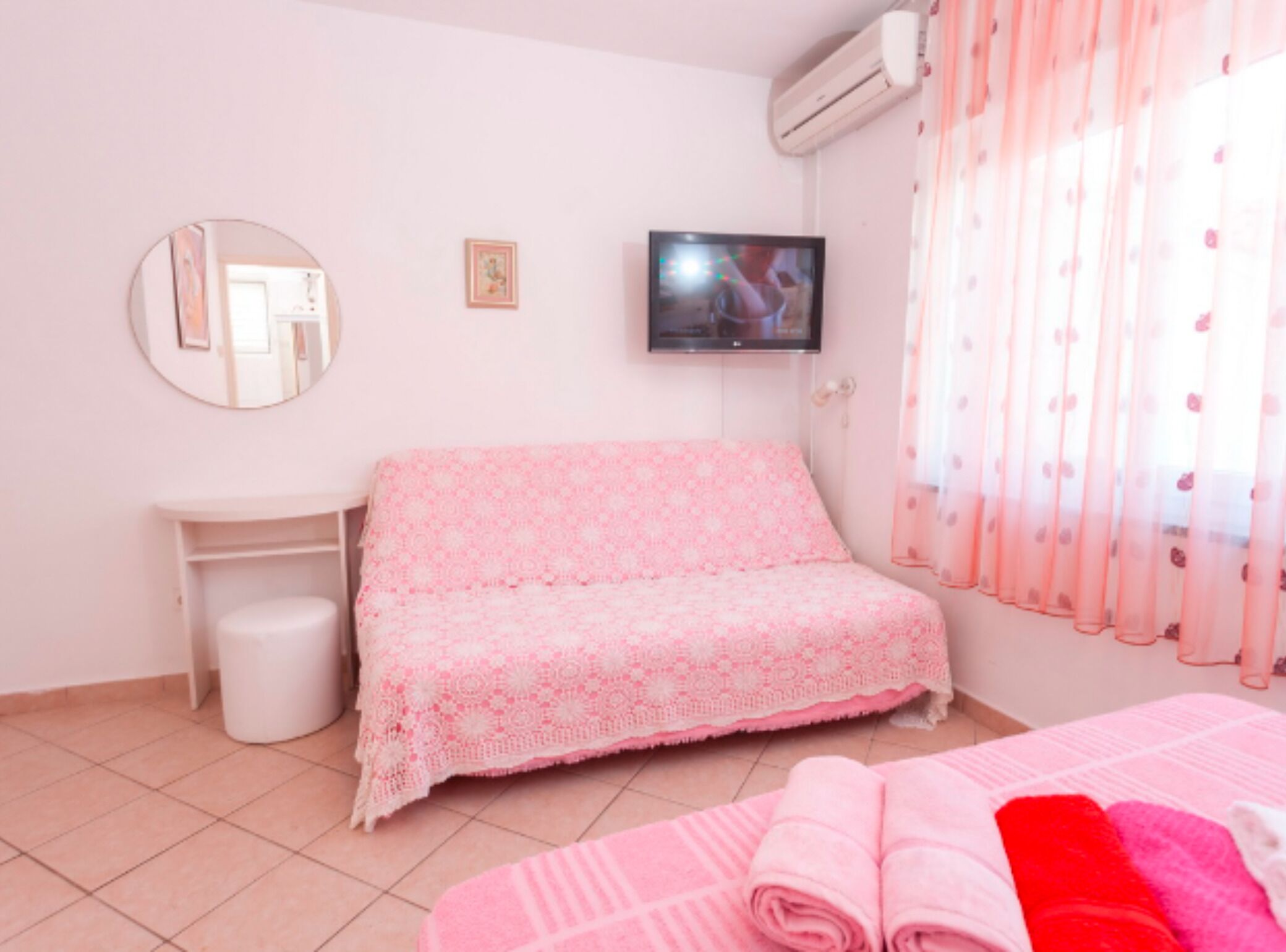 Une chambre avec un lit rose, une petite table de chevet, un miroir, une télévision et une fenêtre avec des rideaux légers.