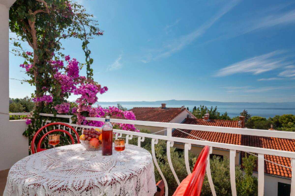 A balcony with a table set for a meal, overlooking a coastal view with houses and a clear sky.
