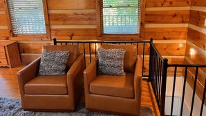 L’intérieur chaleureux d’un chalet avec deux fauteuils en cuir, une petite table en bois et une fenêtre équipée de stores.