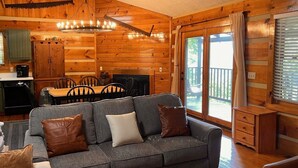 Un intérieur de chalet chaleureux avec un canapé gris, des murs en bois, un coin repas et une vue à travers la porte vitrée.