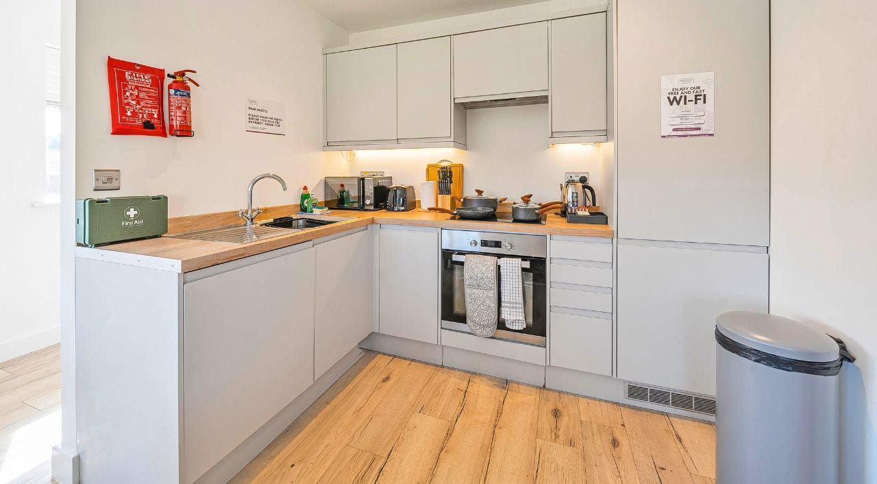 A modern kitchen with white cabinets, a built-in oven, and a stainless steel sink. There is a fire extinguisher and a first aid kit on the counter.