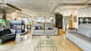 Un vaste salon doté d’une cheminée, de canapés en cuir, d’une table basse en verre et, au fond, d’une cuisine moderne.