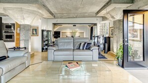 Un salon moderne avec un canapé en cuir gris, une table basse en verre et une cuisine en arrière-plan.