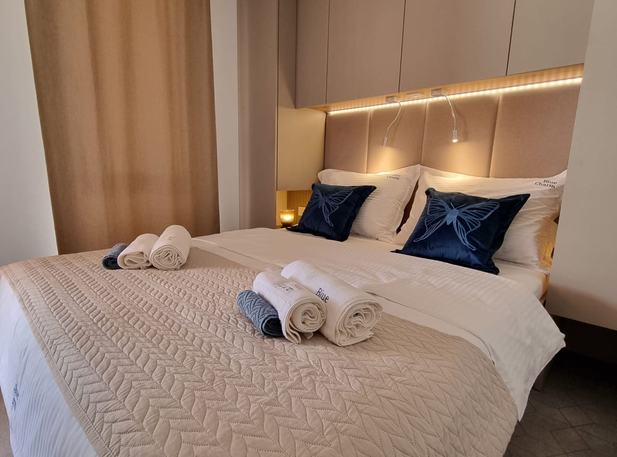 A hotel room with a large bed, beige quilted bedspread, two rolled towels, and a headboard with integrated lighting.