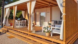 A covered outdoor seating area with wooden flooring, white curtains, and a table with chairs. - Blue Charm Mobile Home (Orebic)