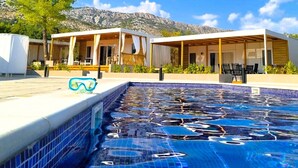A modern pool area with a clear blue sky, a mountain backdrop, and a covered seating area with outdoor furniture. - Blue Charm Mobile Home (Orebic)
