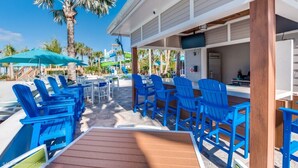A poolside area with blue chairs and a wooden deck, palm trees, and a building with a bar area. - Last Minute Disney! Private Pool, Hot Tub & Themed Bedrooms. Book Now! #108121 (Orlando)