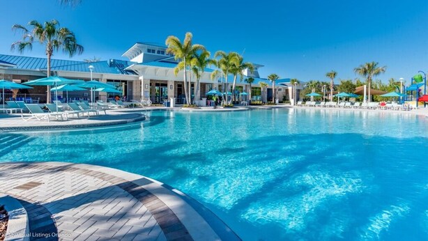 A large swimming pool with lounge chairs and umbrellas, surrounded by palm trees and a modern building with a glass facade. - Last Minute Disney! Private Pool, Hot Tub & Themed Bedrooms. Book Now! #108121 (Orlando)