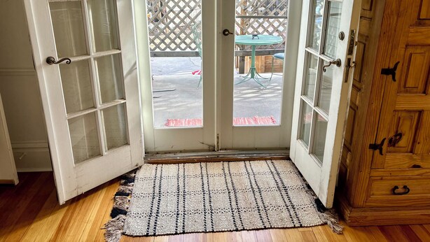 Une porte en bois avec des panneaux de verre, un paillasson en fibres tressées et une vue sur un patio avec une table et des chaises.
