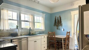 Une cuisine équipée d’un évier, d’une cuisinière et de placards, une table à manger avec des chaises, et une fenêtre avec des rideaux.