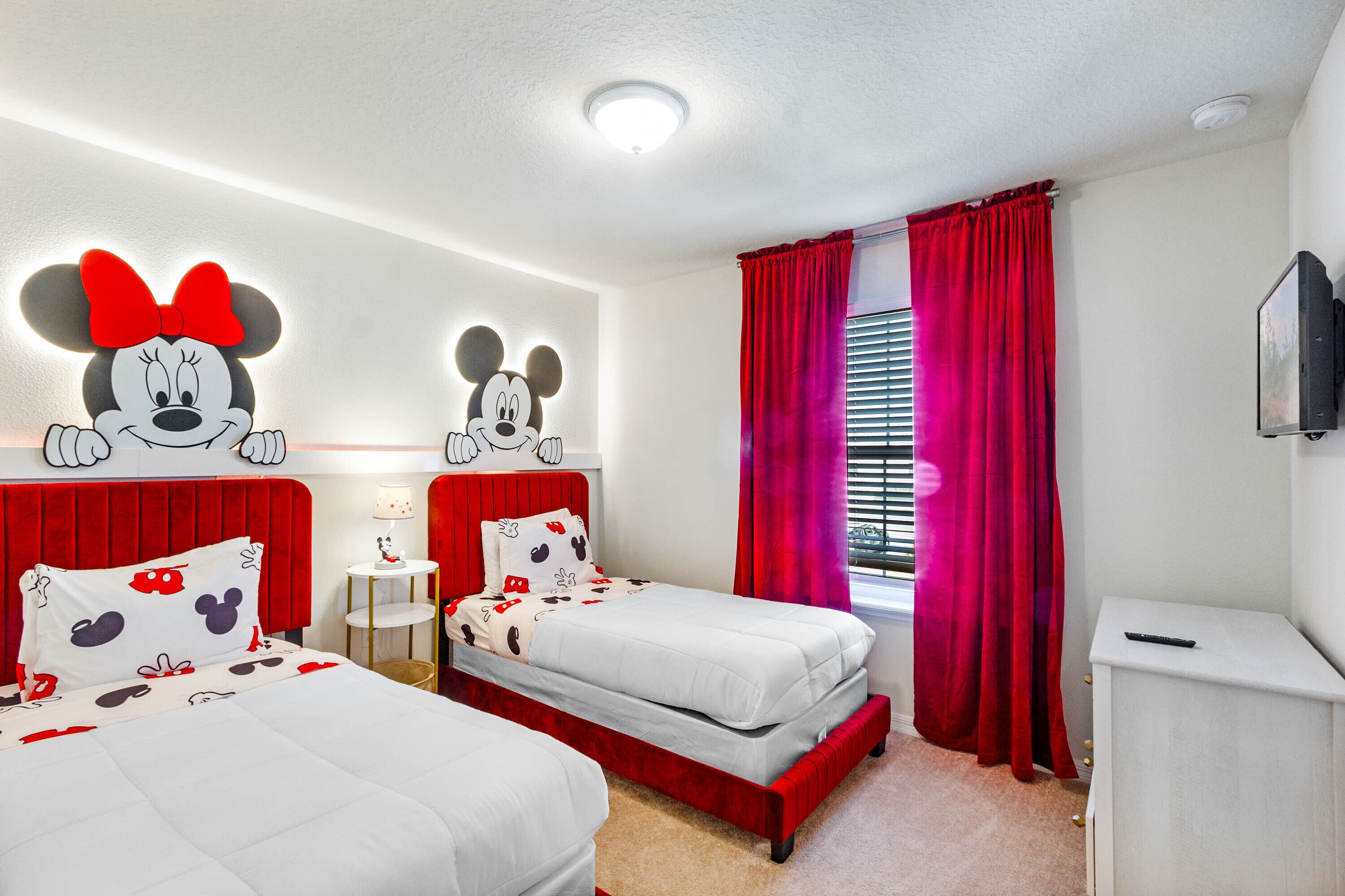 A hotel room with two beds, red curtains, and Mickey Mouse wall decorations.