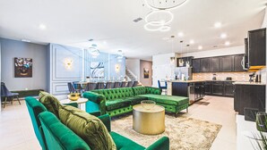 A modern living room with a green sectional sofa, a dining area with a long table and chairs, and a kitchen with dark cabinets and stainless steel appliances.
