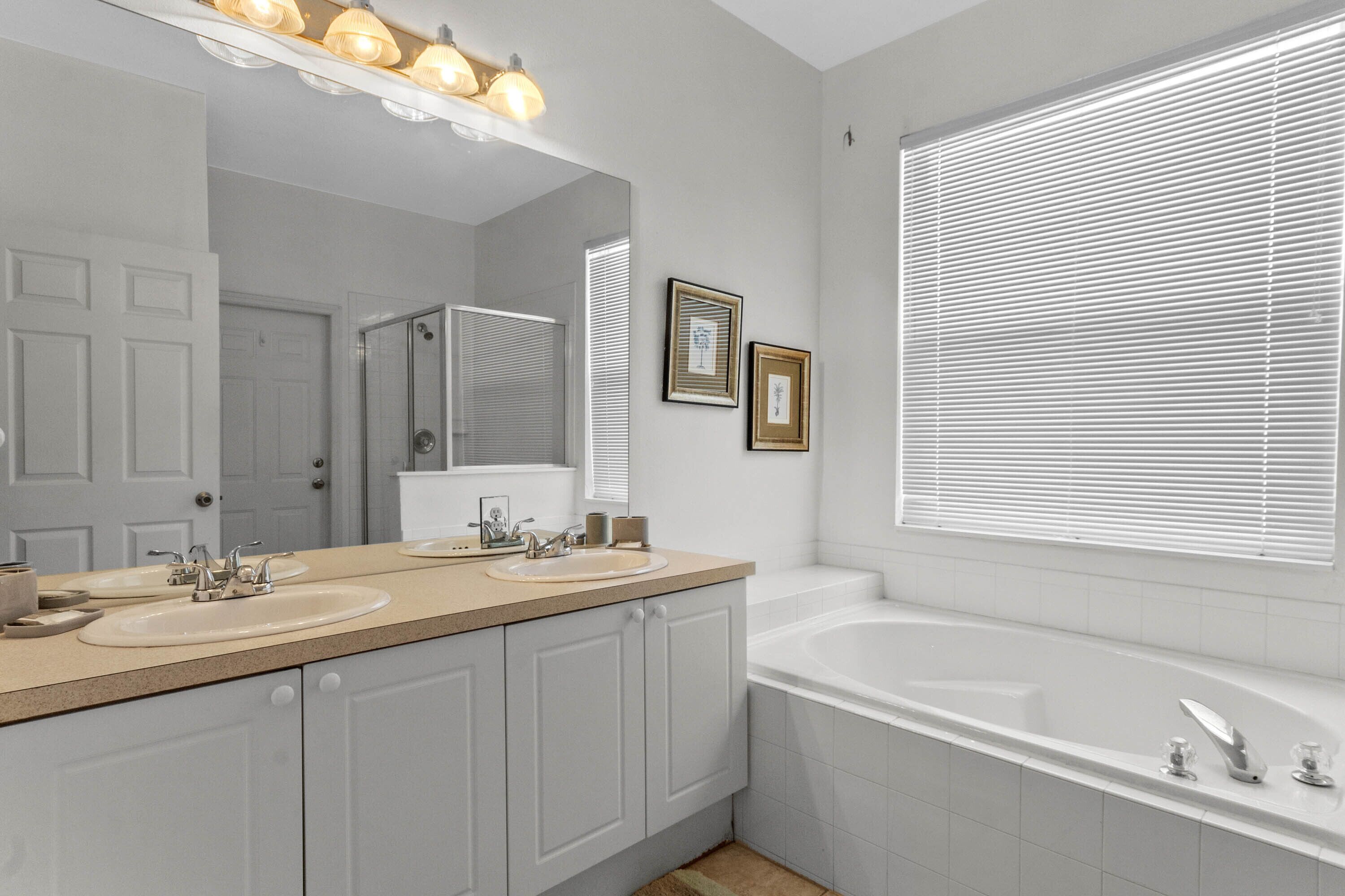 A modern bathroom with a double sink vanity, a large mirror, a bathtub, and a window with blinds.