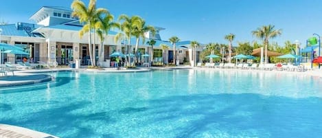A large outdoor swimming pool with palm trees and umbrellas, surrounded by a modern building with a blue roof.