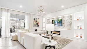 A modern living room with a sofa, a coffee table, a fireplace, a TV mounted on a marble wall, and a balcony with outdoor furniture. - Last Minute Disney! Private Pool, Hot Tub, Game Room & Themed Bedrooms. Book Now! #111505 (Orlando)