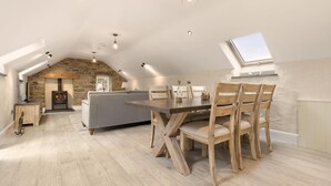Een knus loft-achtig vertrek met een stenen muur, een open haard, een eettafel met houten stoelen en een dakraam boven de tafel.