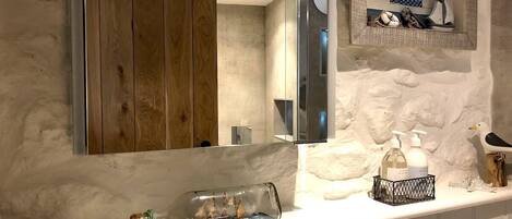 Une salle de bain avec un meuble-lavabo en bois, un grand miroir, une petite étagère ornée d’objets décoratifs et un bocal en verre contenant une maquette de bateau.