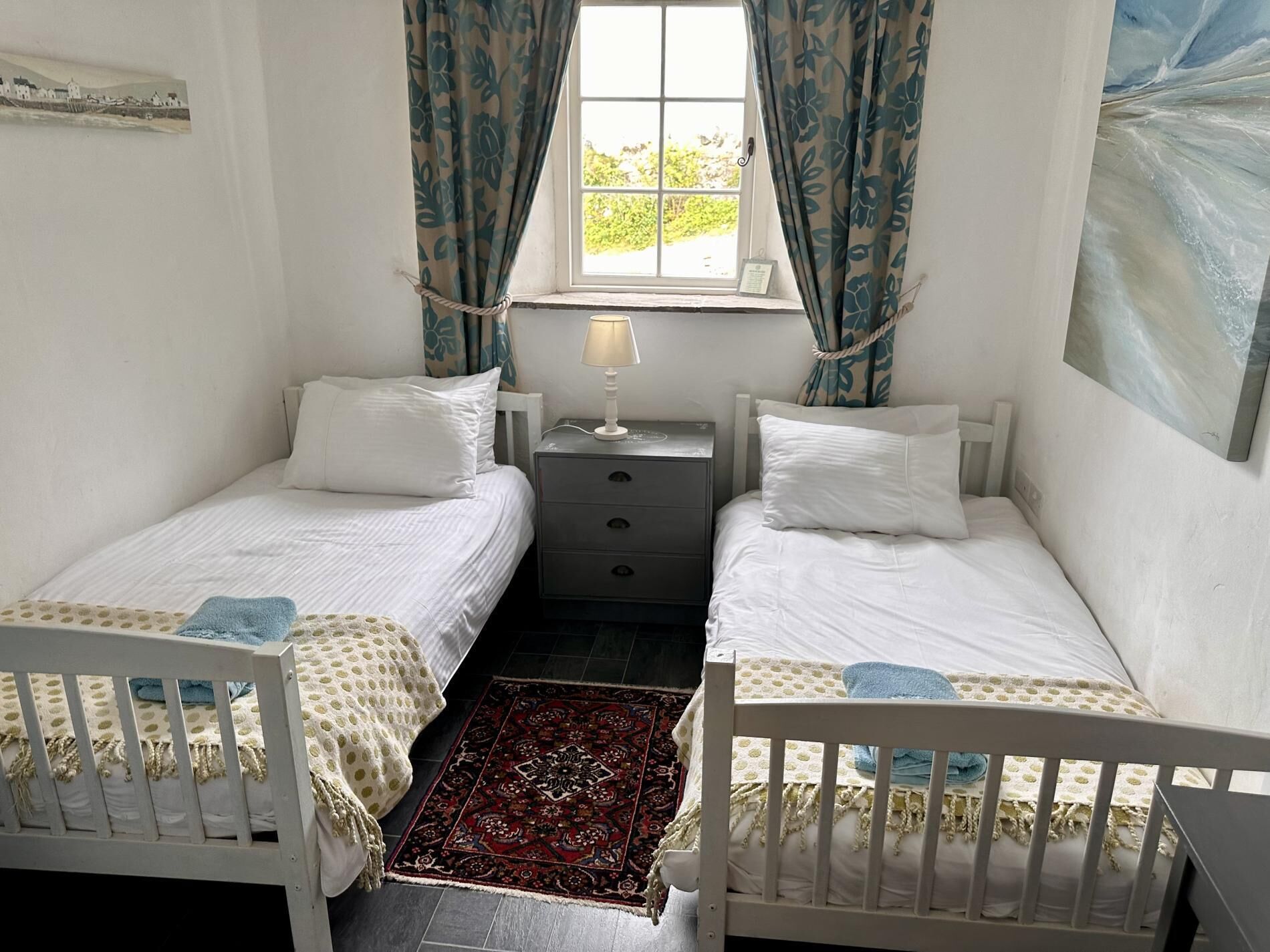 Une chambre avec deux lits simples, une table de chevet avec une lampe, un petit tapis et une fenêtre avec des rideaux.