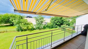 Ein Balkon mit gelbem Streifenmarkisenstoff, einem Metallgeländer und Blick auf eine grüne Rasenfläche und Bäume.