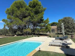 Une piscine entourée d’arbres et d’un bâtiment en pierre, avec des chaises longues et un parasol à proximité.