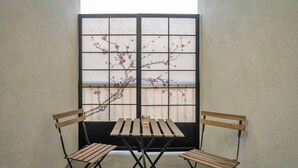 Une pièce avec une petite table et deux chaises en bois, une fenêtre à cadre noir ornée d’un motif floral et un mur uni.