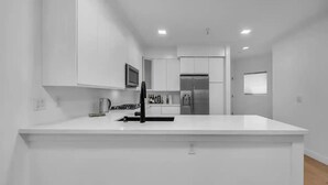 Une cuisine moderne avec des meubles blancs, un îlot central, des appareils en acier inoxydable et des spots encastrés au plafond.