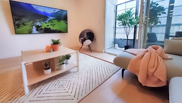 Un salon moderne doté d’un grand téléviseur à écran plat, d’une table basse blanche agrémentée de plantes, d’un canapé recouvert d’un plaid et d’une grande fenêtre offrant une vue sur l’extérieur.