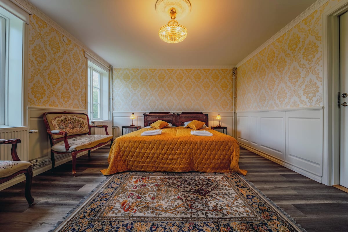 A bedroom with a large bed, a wooden chair, a patterned rug, and a chandelier.