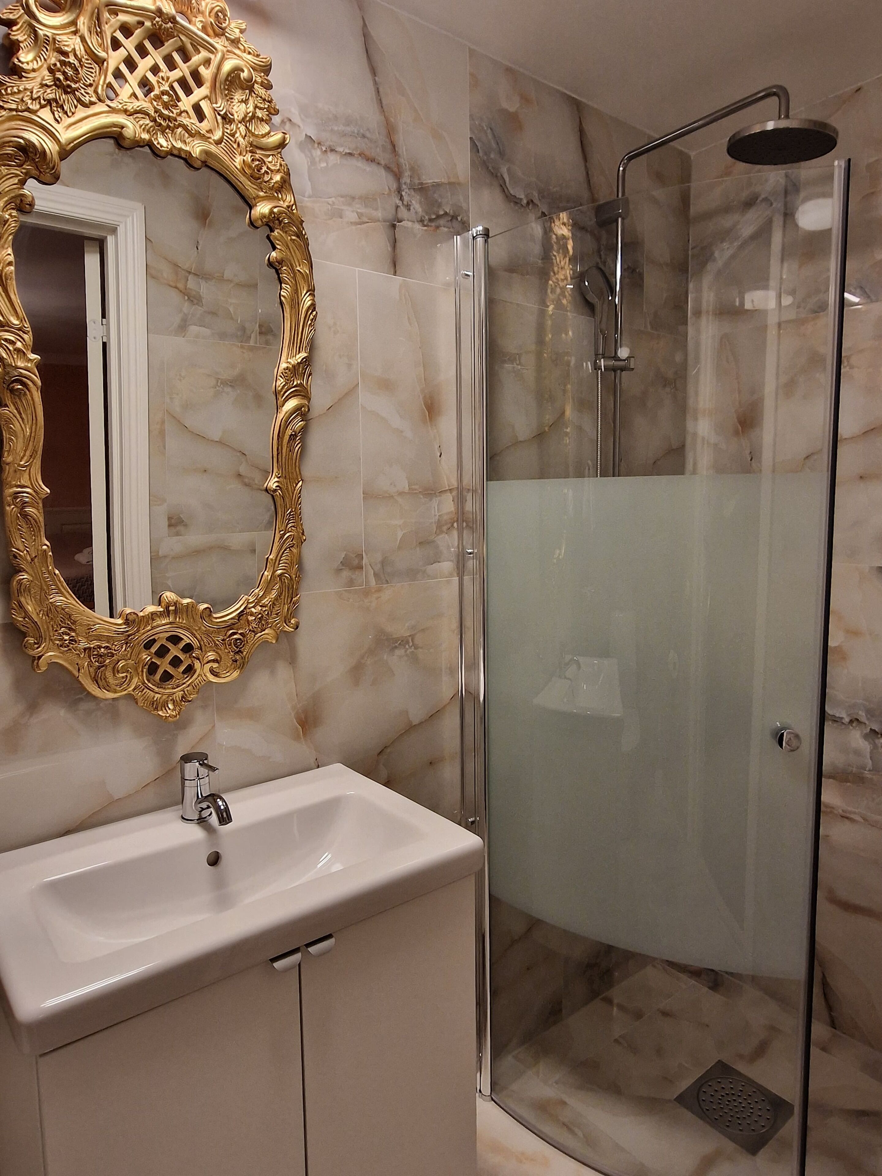 A bathroom with a marble wall, a gold-framed mirror, a white sink, and a glass-enclosed shower.