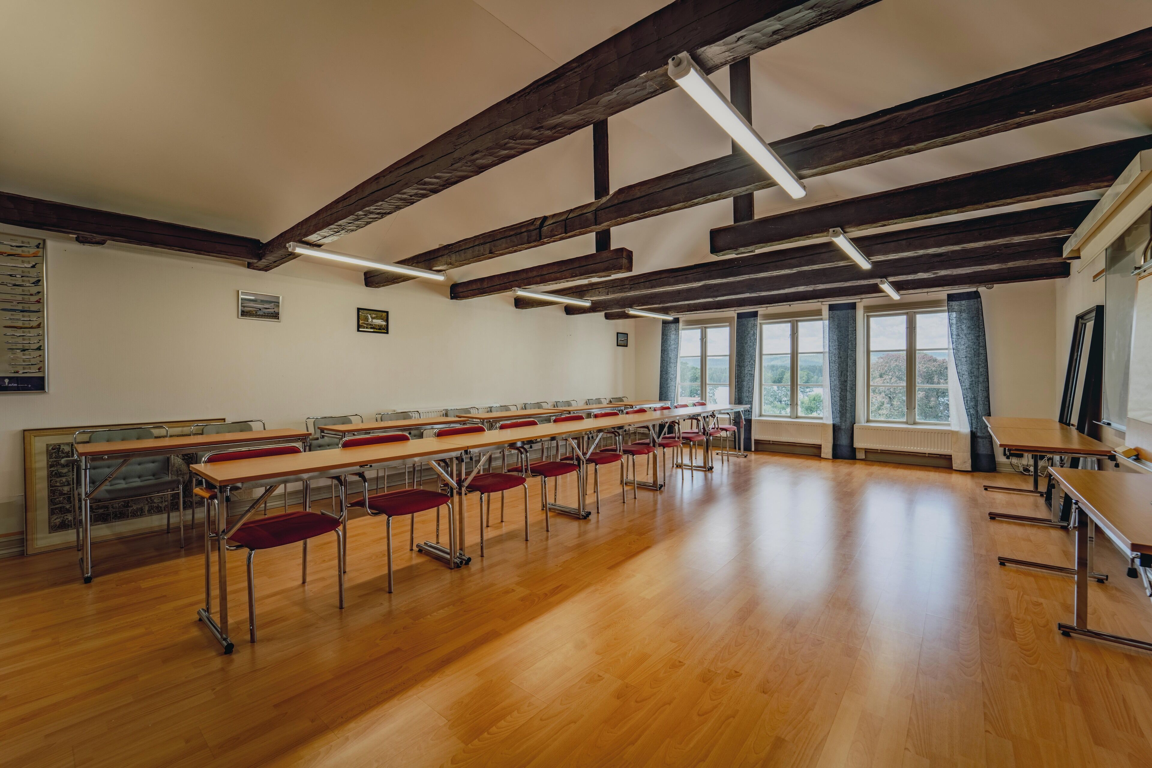 A classroom with wooden desks and chairs, large windows, and exposed wooden beams on the ceiling.